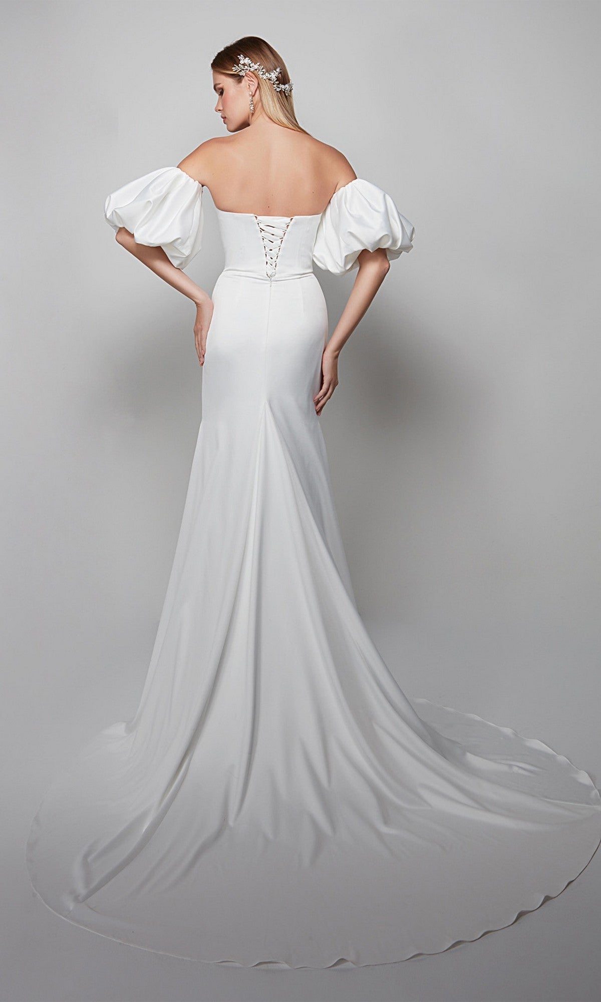 A woman wearing the Alyce 7056 long formal dress with puff sleeves, a lace-up back, and an off-the-shoulder sweetheart neckline stands on a gray background, facing away from the camera.